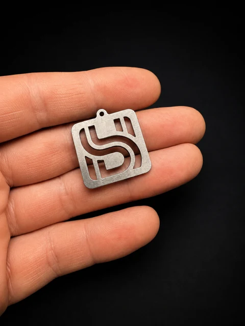 A small square metal part with a cut-out letter design resting on a hand