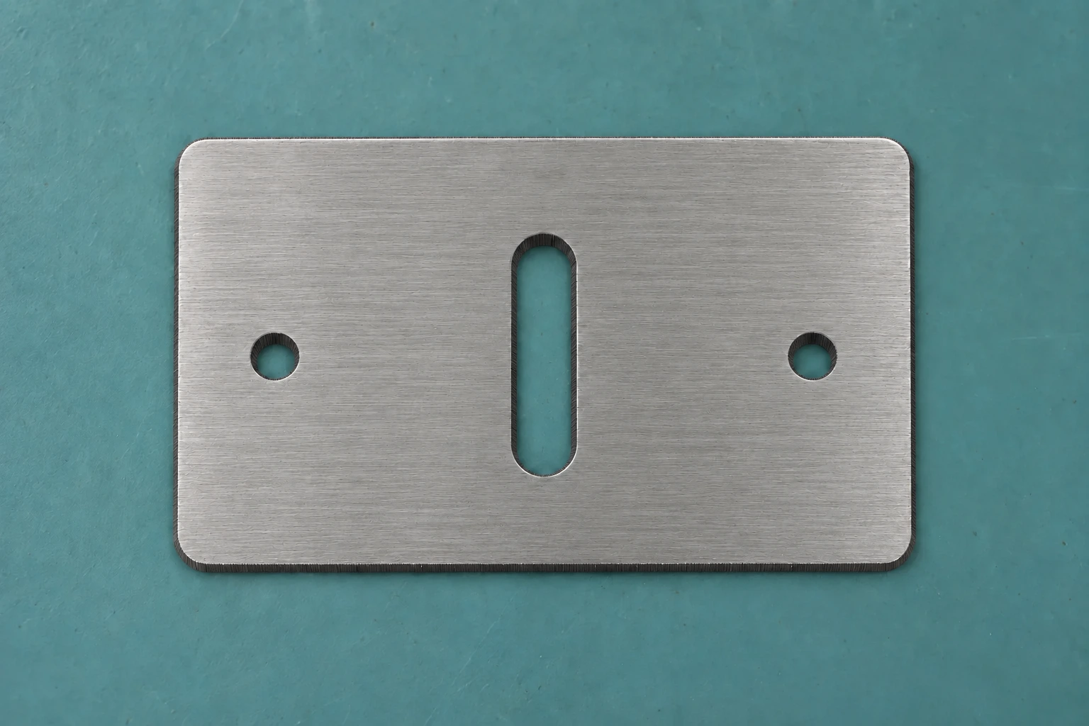 Suitable upload photo: a fresh flat steel plate with a uniform lengthwise brushed finish viewed from above on a plain blue-green surface, with both holes and the central slot clearly visible